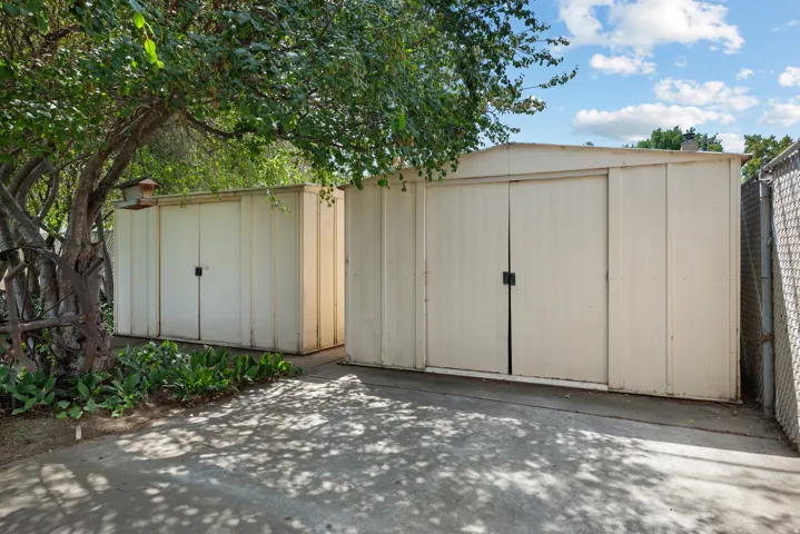 Two metal storage sheds with double doors, concrete pad, and surrounding mature trees