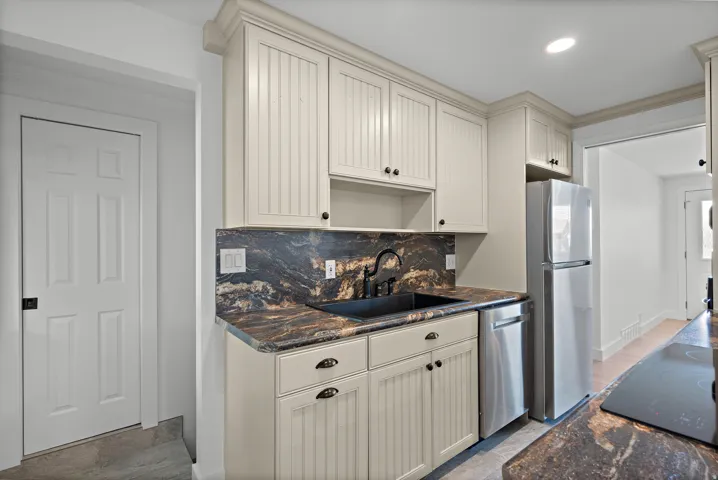 Cream-painted shaker-style cabinetry with black hardware, dark stone countertops and backsplash, black undermount sink with a matte black faucet, and stainless steel appliances