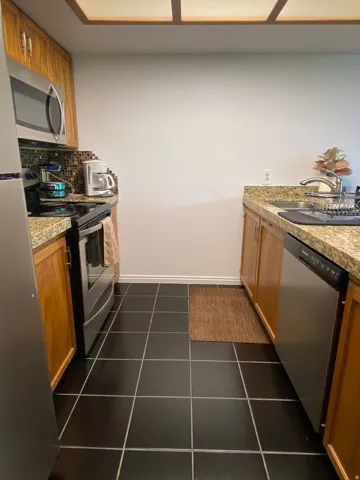 Kitchen featuring wood cabinetry, stainless steel appliances, speckled countertops, a mosaic tile backsplash, and black tile flooring