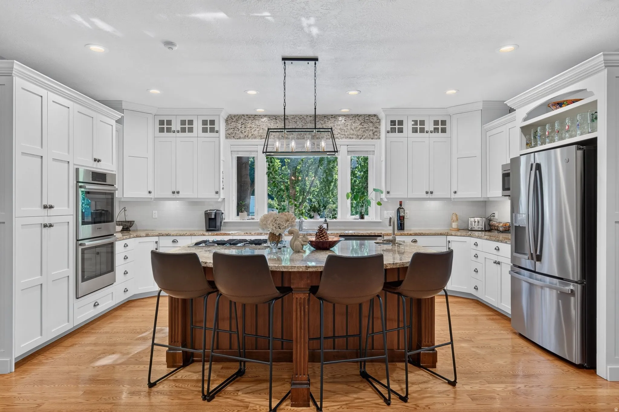 White shaker cabinetry, stainless steel double ovens, wood-finish flooring, central island with seating, and a mosaic tile backsplash