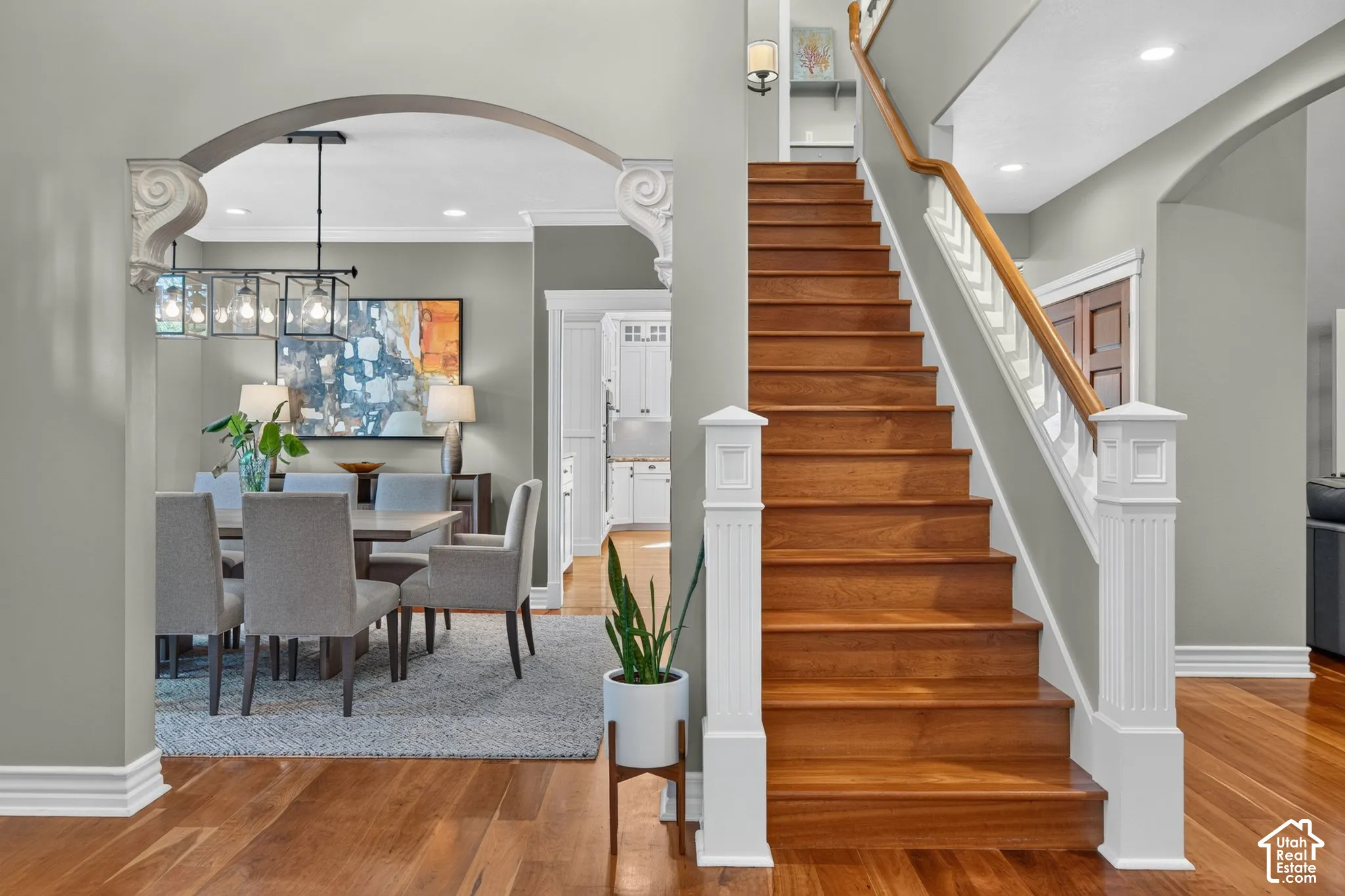 Hardwood flooring, wood staircase with white newel posts, dining area with a linear chandelier, and arched doorways with corbel details