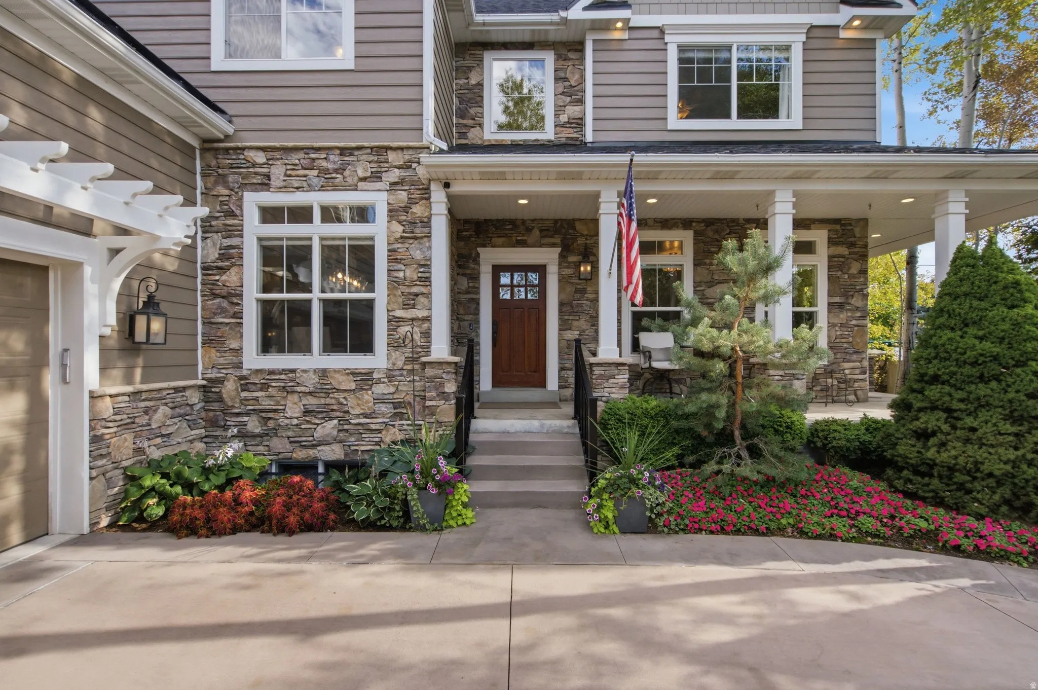 Stone facade with siding accents, covered front porch, white pergola, concrete driveway, and mature landscaping