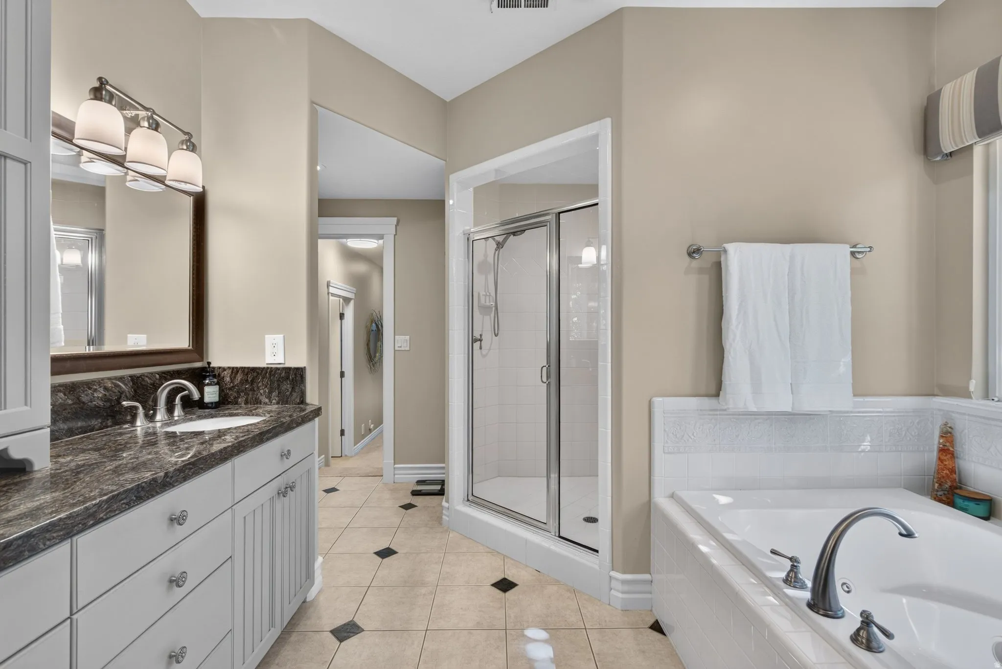 Dual sink vanity with dark countertop, built-in soaking tub, glass-enclosed shower, and tile flooring