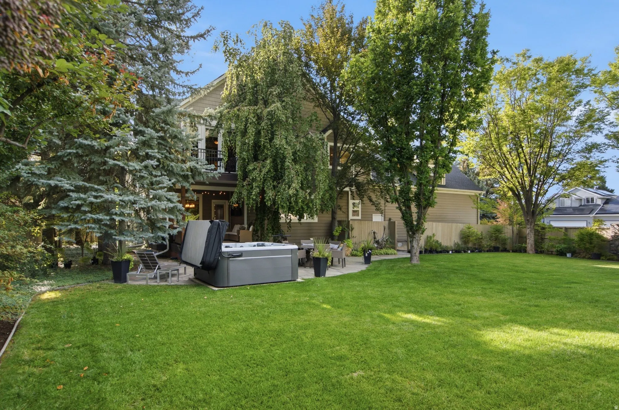 Expansive green lawn with mature trees, a stone patio, and a deck with a black metal railing