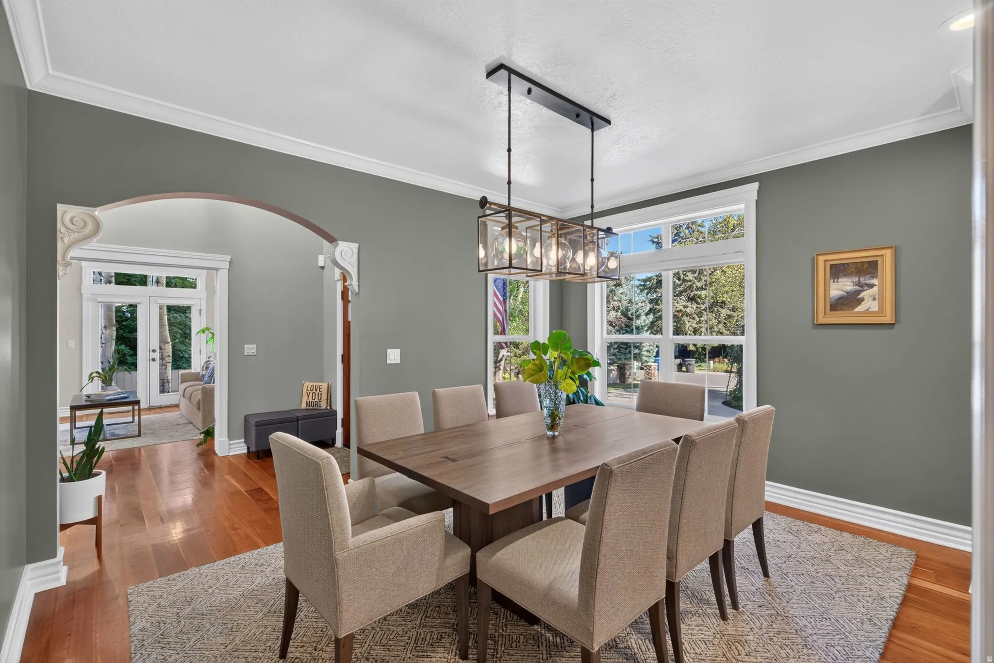 Hardwood flooring, arched doorway with decorative corbels, white crown molding, large multi-pane windows, and a rectangular chandelier with clear glass panels