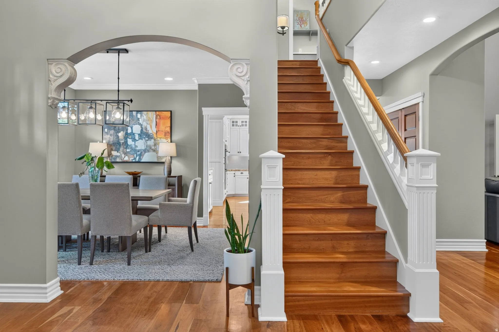 Hardwood flooring, wood staircase with white newel posts, dining area with a linear chandelier, and arched doorways with corbel details