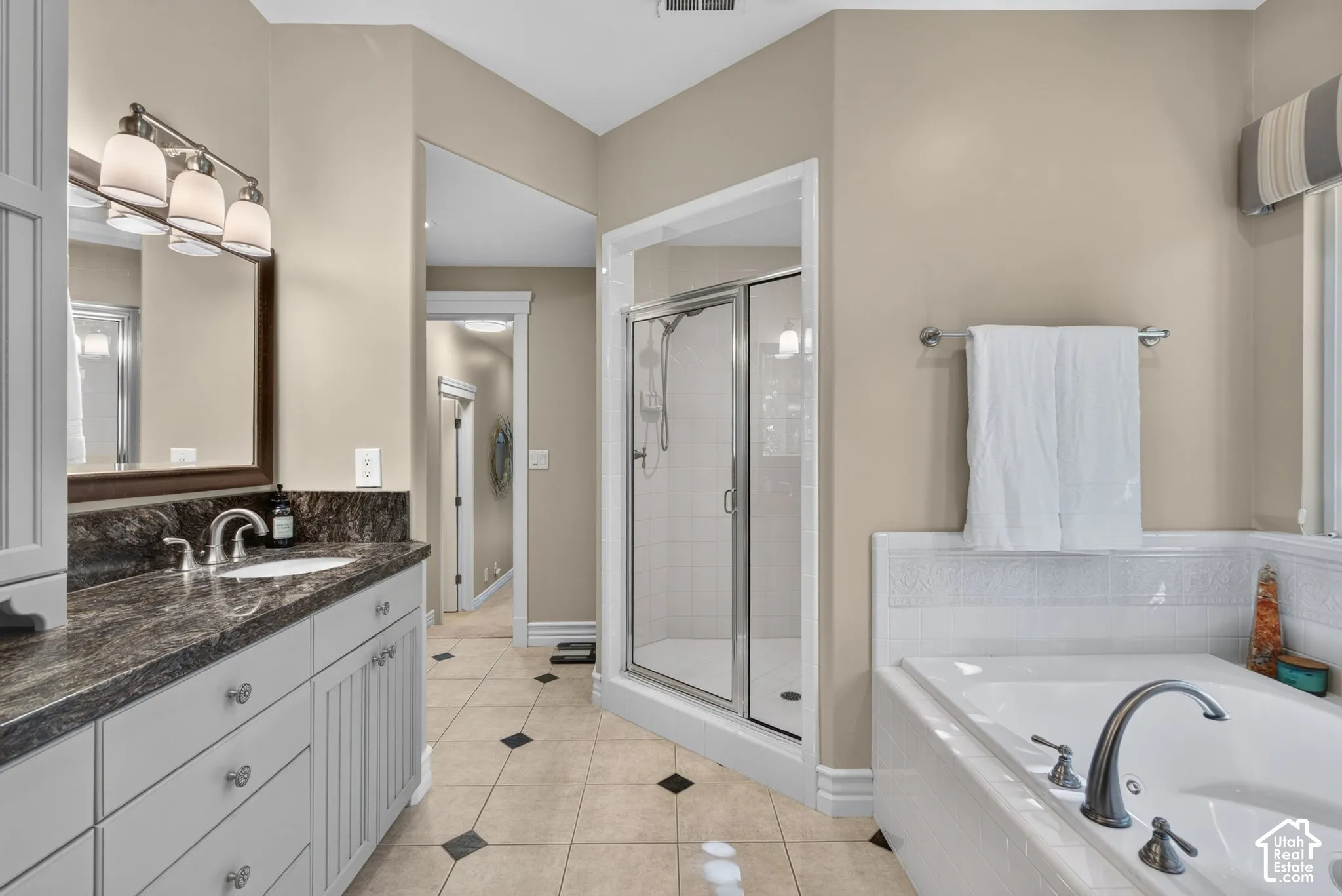 Dual sink vanity with dark countertop, built-in soaking tub, glass-enclosed shower, and tile flooring