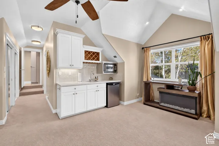 Vaulted ceiling featuring a ceiling fan, built-in wet bar with white cabinetry and wine rack, large window with natural light, and plush carpeting