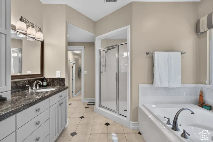 Dual sink vanity with dark countertop, built-in soaking tub, glass-enclosed shower, and tile flooring