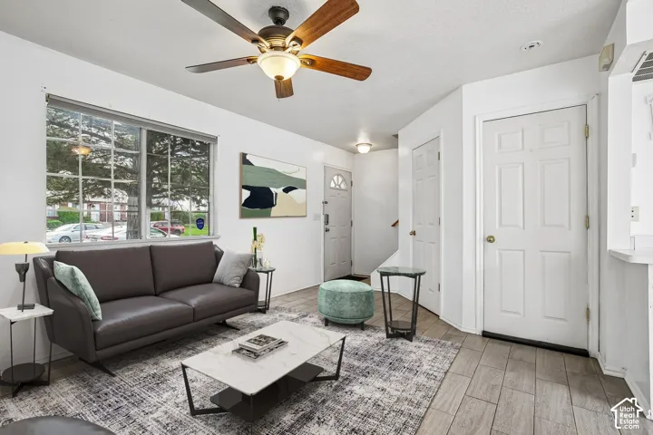 Bright living area featuring large multi-pane windows with natural light, dark upholstered seating, wood-finish flooring throughout, ceiling fan with integrated lighting, and neutral wall finishes with white trim and doors