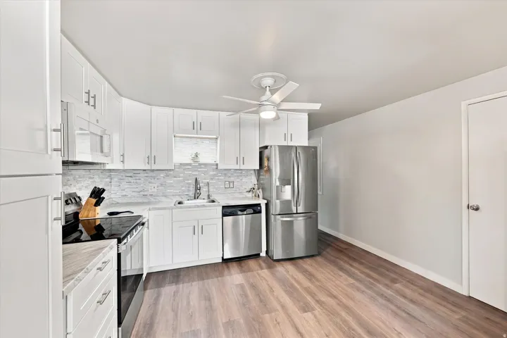 White shaker-style cabinetry, stainless steel appliances, wood-finish flooring, mosaic tile backsplash, and a ceiling fan with integrated lighting. Entrance door off kitchen to access back yard and ADU.