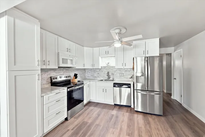 White shaker-style cabinetry, stainless steel appliances, light-toned countertops, tile backsplash, and wood-finish flooring