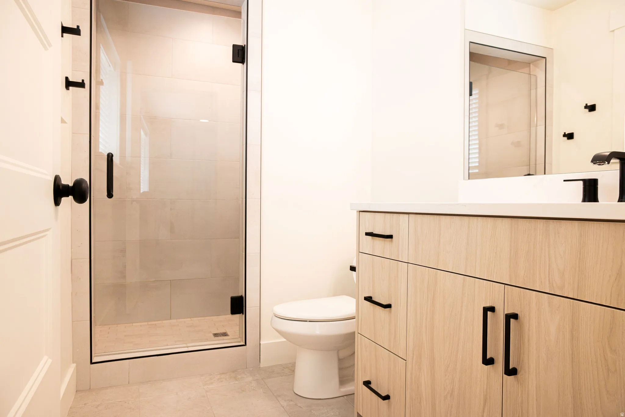 Glass-enclosed shower with black trim, light wood-finish vanity with black hardware, white countertop, and tiled flooring