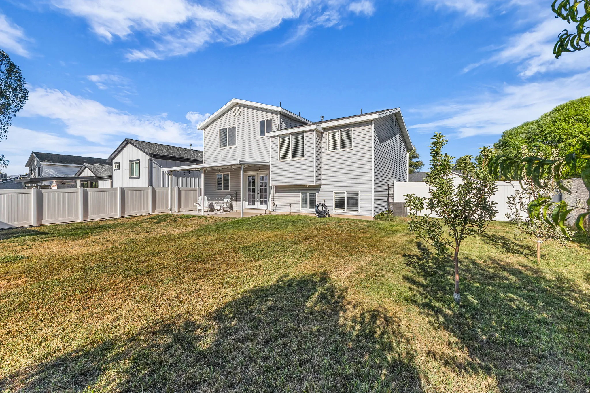Expansive lawn area with a two-story exterior, covered patio, white vinyl fencing, and mature plantings