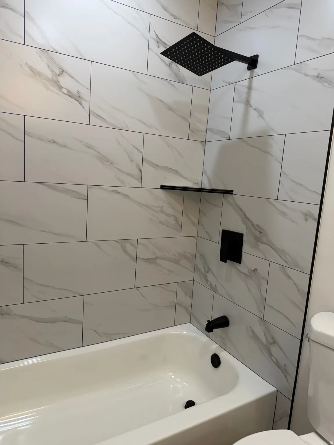 Bathtub with white subway tile surround featuring gray veining, matte black rain shower head, matte black shower valve, and matte black shelf