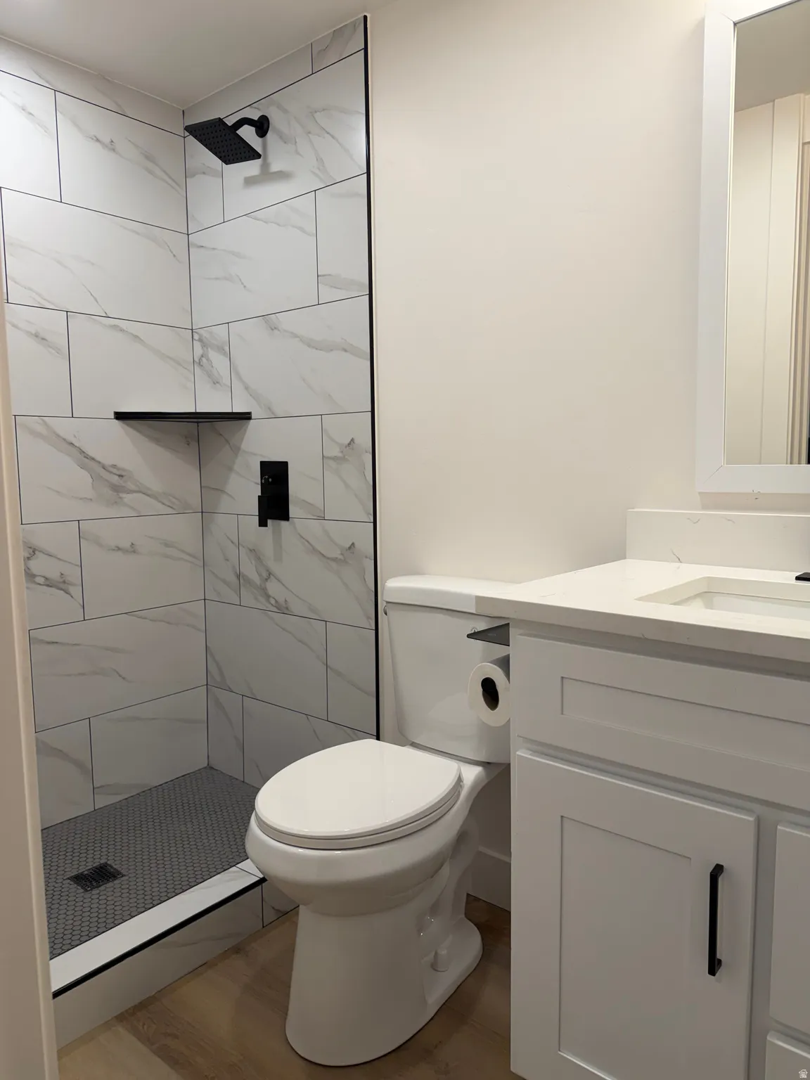 Walk-in shower with large format tile surround and hexagonal floor tile, matte black shower fixtures, single vanity with white shaker cabinetry, and wood-finish flooring