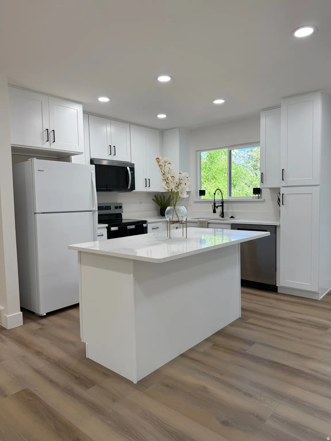 Bright kitchen featuring recessed lighting, white cabinetry with black hardware, a center island with a stone countertop, wood-finish flooring, and a large window