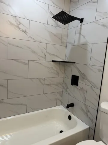 Bathtub with white subway tile surround featuring gray veining, matte black rain shower head, matte black shower valve, and matte black shelf
