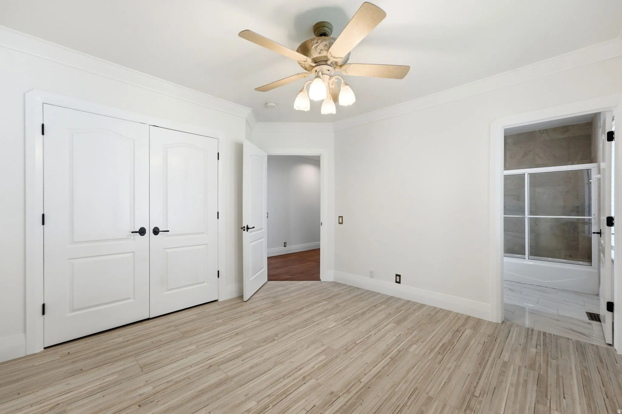 Wood-finish flooring, ceiling fan with light fixture, double panel doors with matte black hardware, crown molding, and an attached bathroom with a shower-tub combination