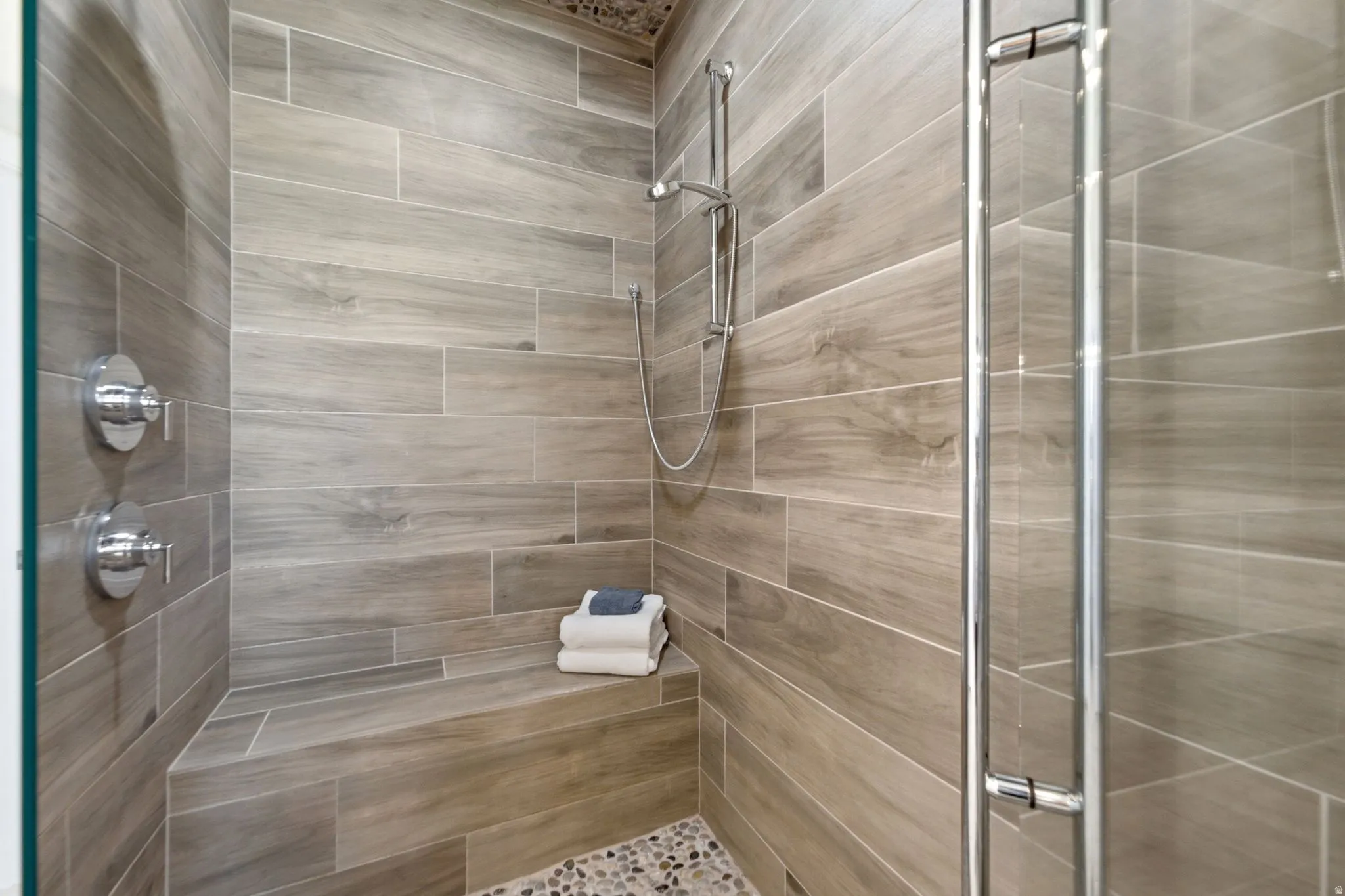 Enclosed shower featuring wood-look tile walls, a built-in bench, a handheld shower fixture, and a mosaic pebble floor