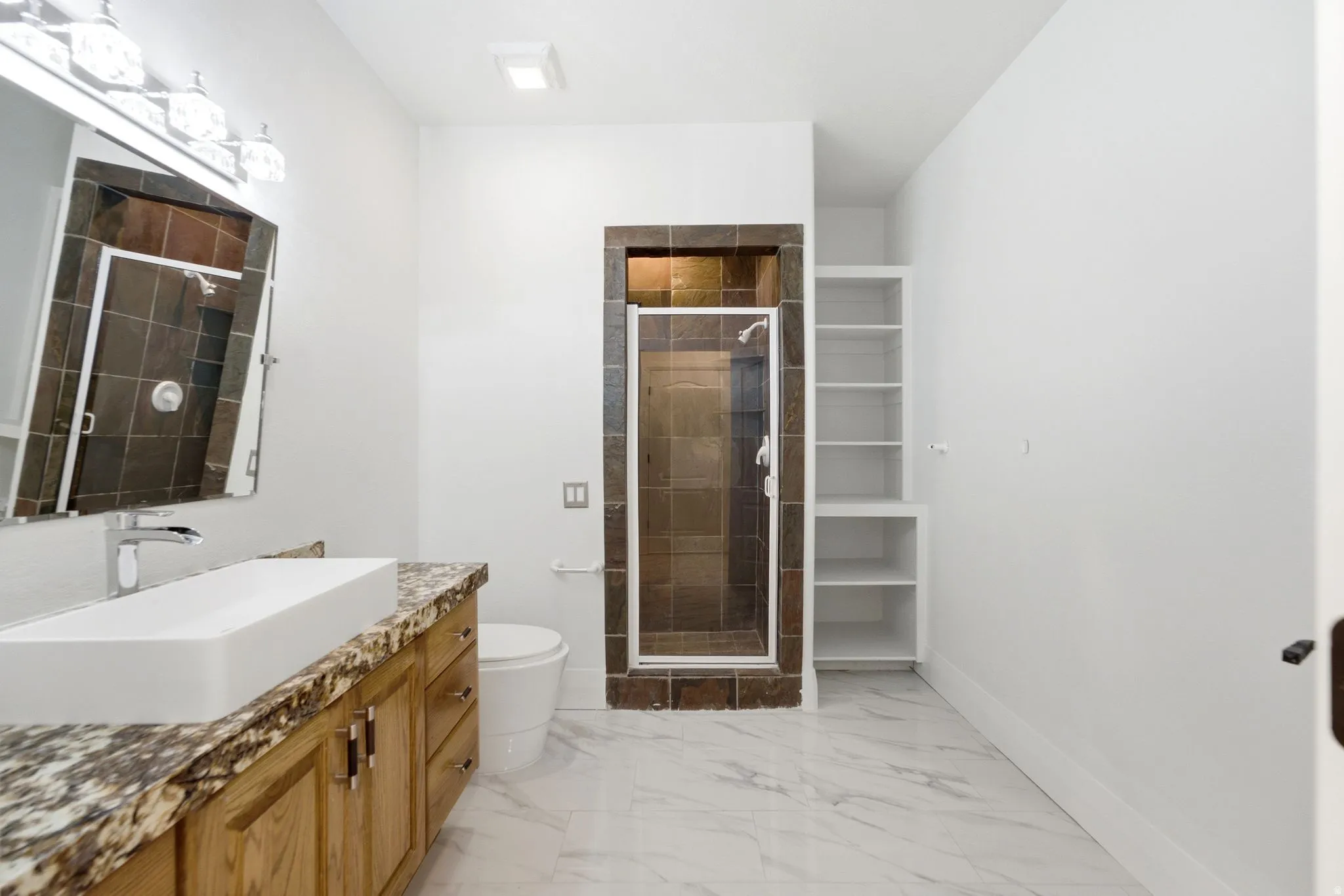 Spacious bathroom featuring a vessel sink, wood-finish vanity with stone countertop, walk-in shower with tiled surround, and integrated shelving