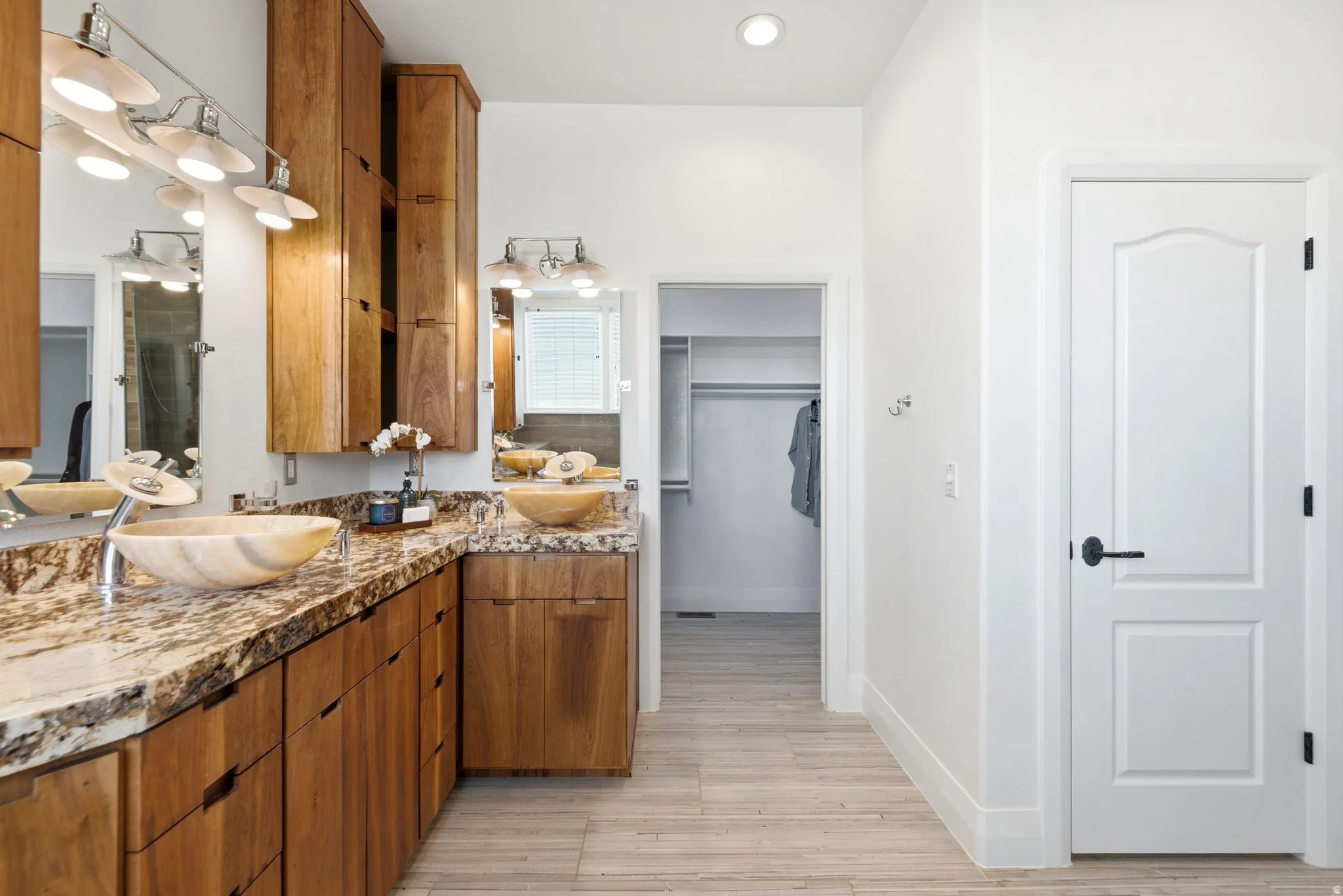 Dual vessel sinks with modern faucets, extensive wood cabinetry, stone-finish countertops, wall-mounted light fixtures, and light-toned flooring