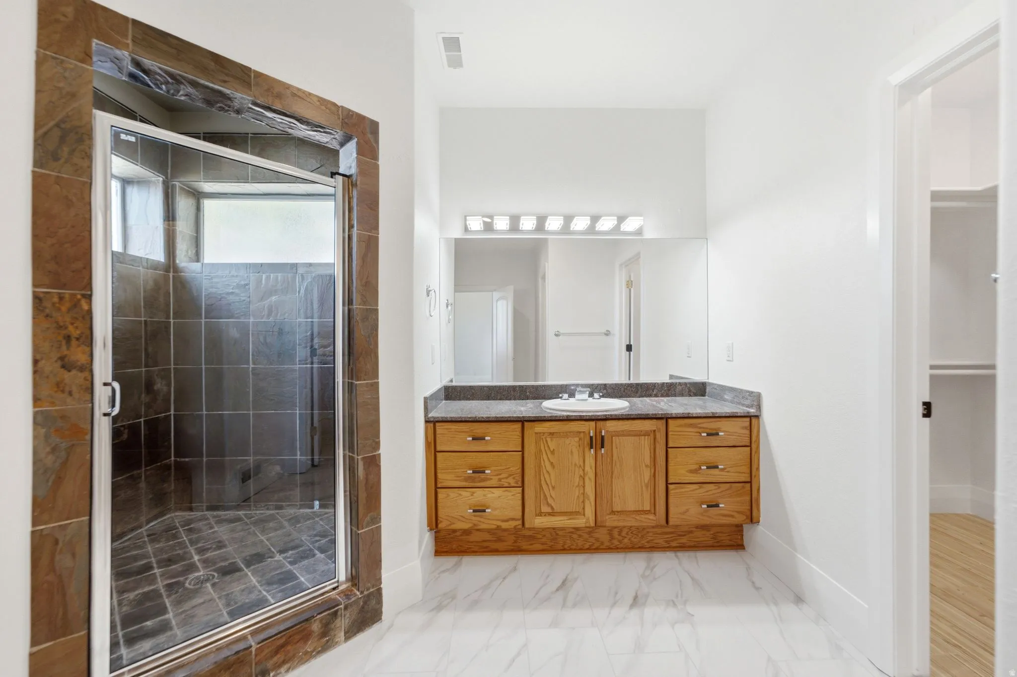 Spacious bathroom featuring a glass-enclosed shower with stone tile, a single vanity with wood cabinetry, a built-in mirror, and white tile flooring