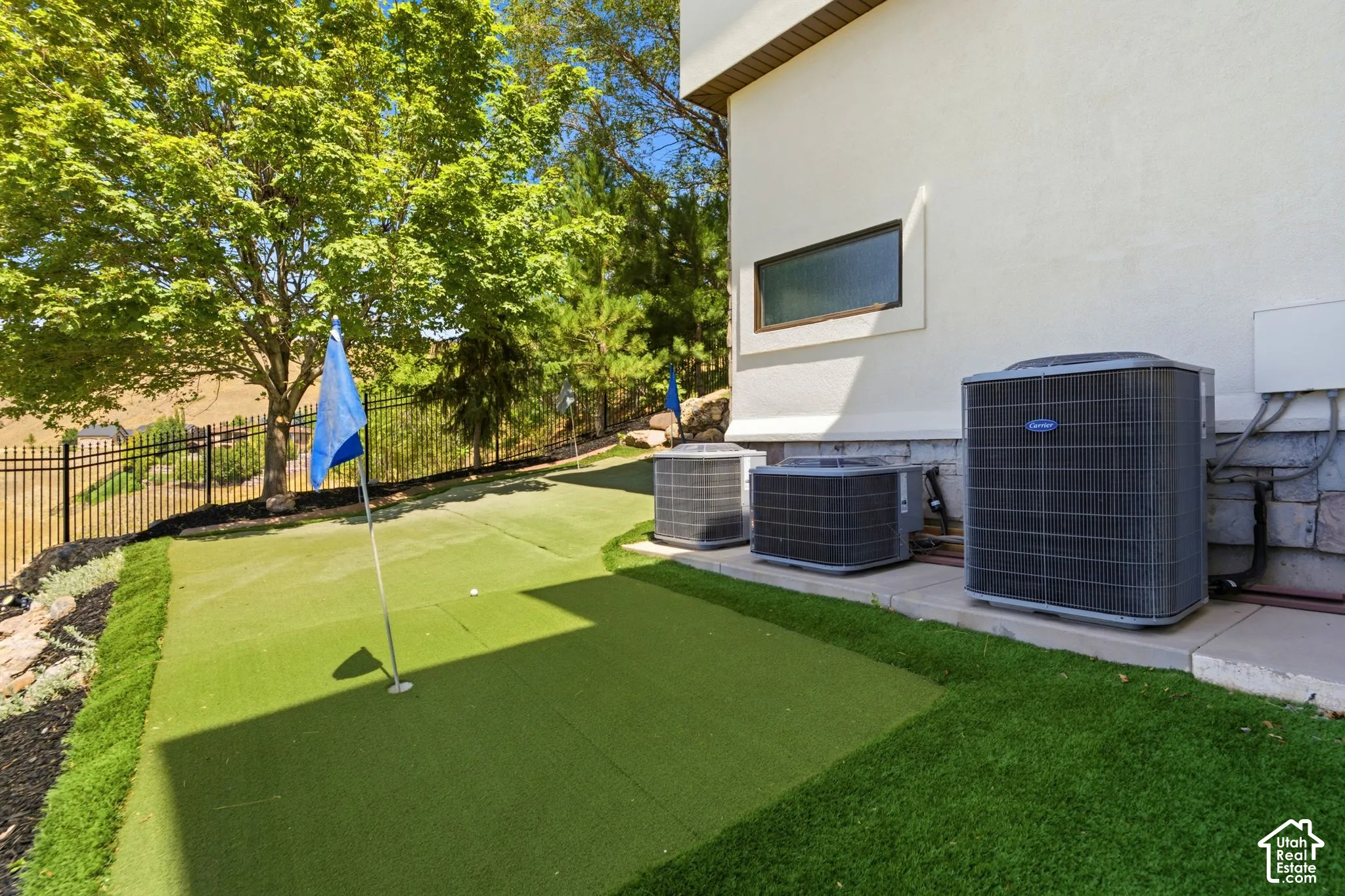 Putting green with artificial turf, bordered by landscaping and a black metal fence
