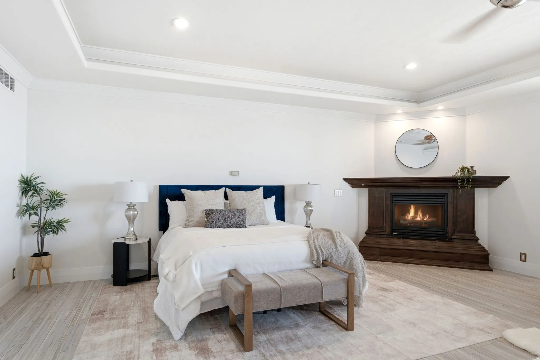Tray ceiling with recessed lighting, wood-finish flooring, corner fireplace with dark wood surround, and wall-mounted ceiling fan