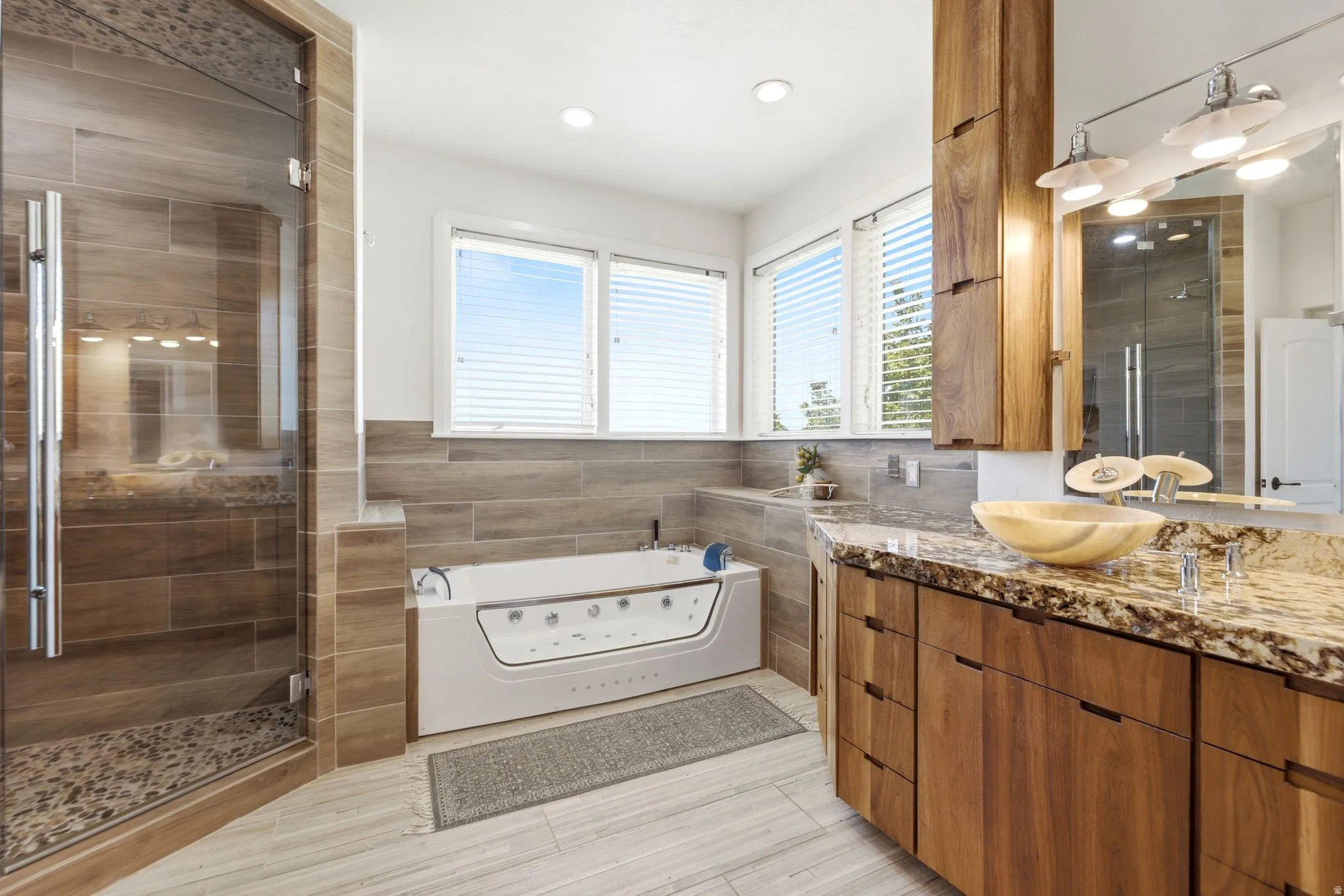 Glass-enclosed shower with tiled walls and pebble floor, soaking tub, wood-finish vanity with stone countertop, and vessel sink