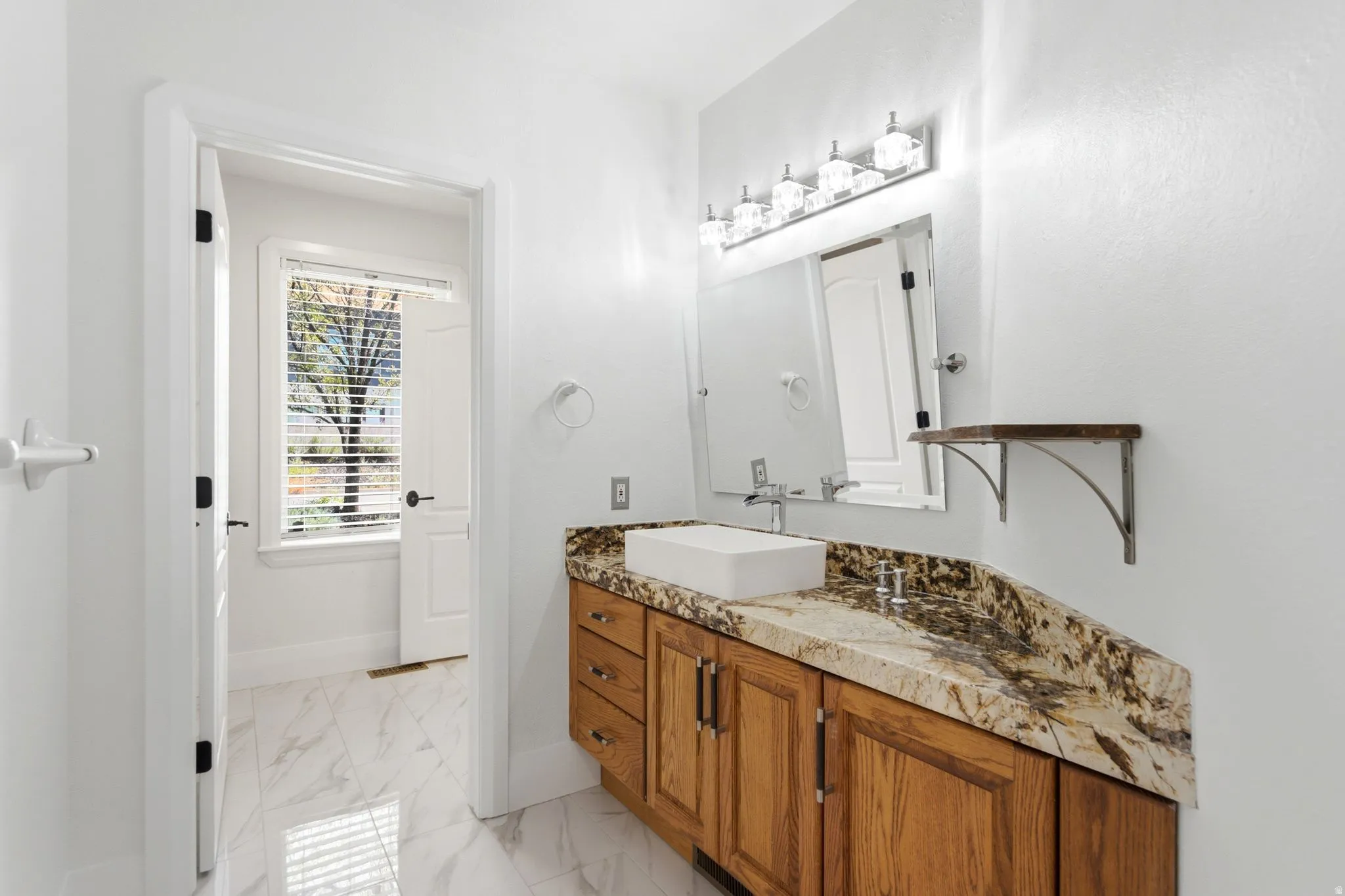 White tile flooring, wood-finish vanity with drawer storage, vessel sink, and a stone-finish countertop
