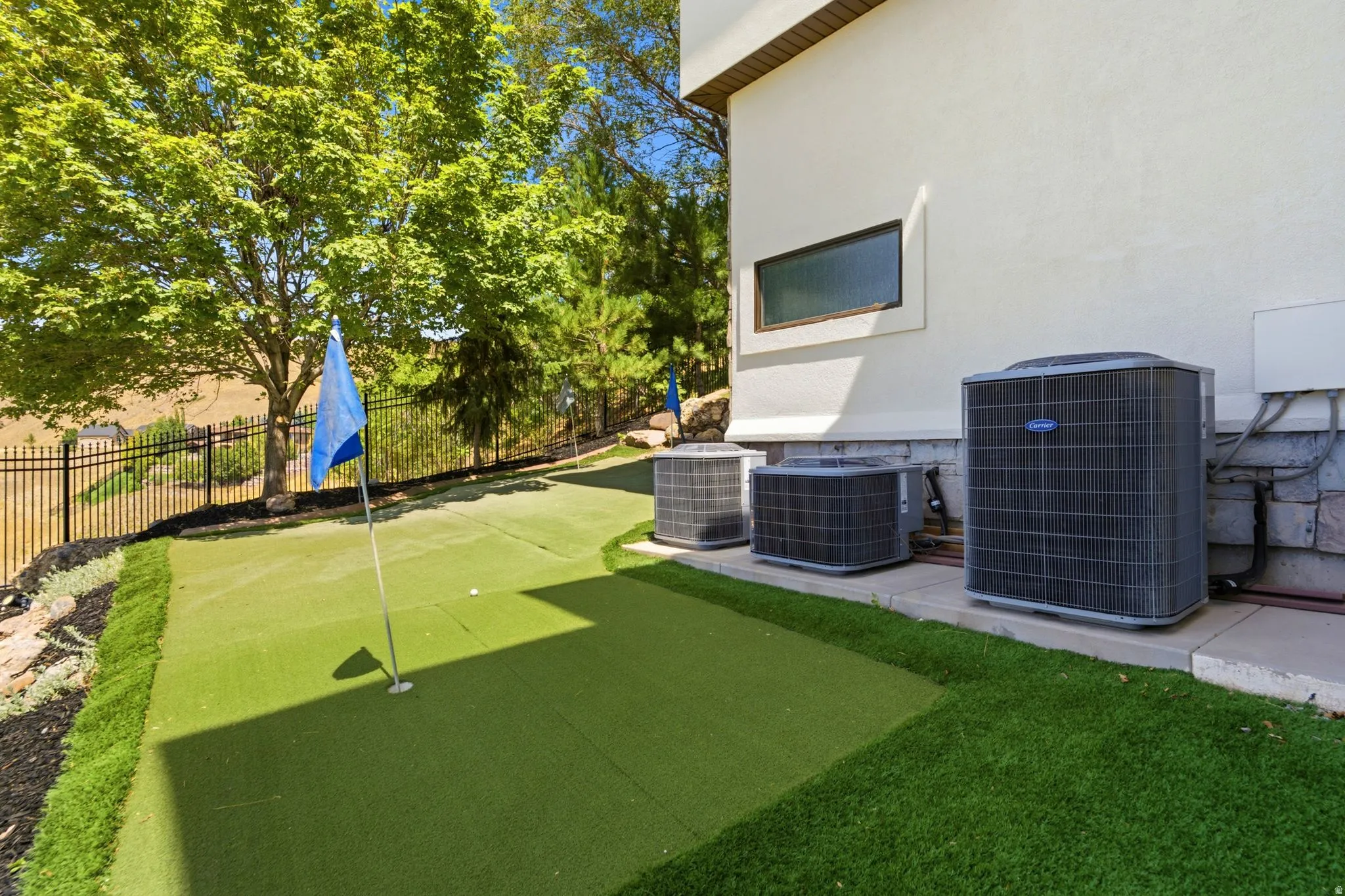 Putting green with artificial turf, bordered by landscaping and a black metal fence