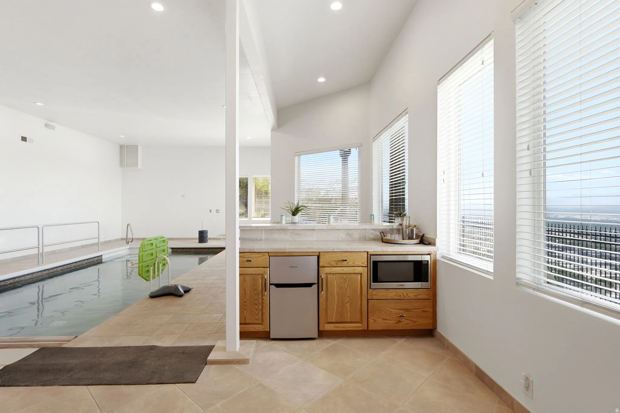 Indoor swimming pool with handrails, tiled pool deck, built-in cabinetry with a microwave, and multiple windows
