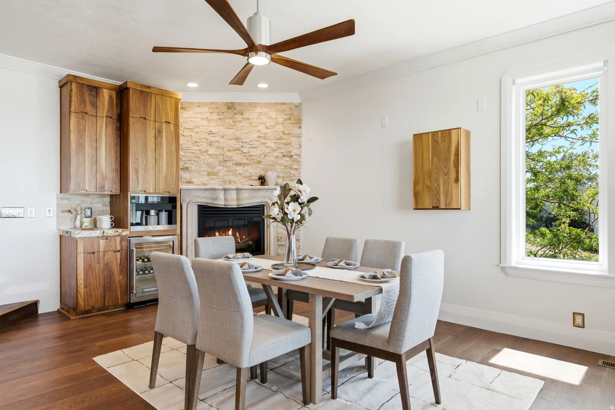 Wood-finish flooring, built-in cabinetry with integrated beverage cooler, stone-clad fireplace with mantel, ceiling fan, and large window