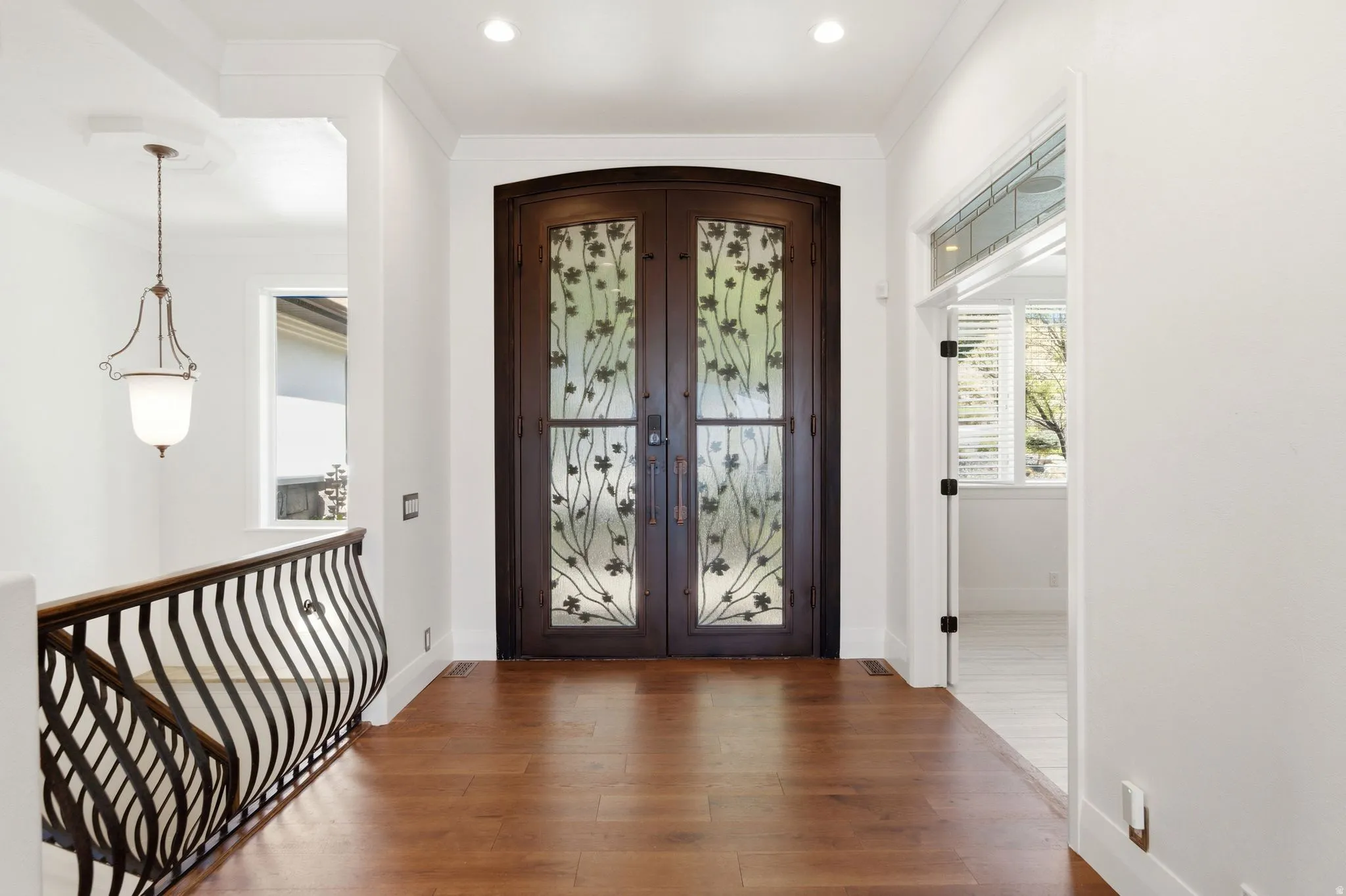 Arched entryway doors with decorative glass panels, wood-finish flooring, wrought iron railing, and recessed lighting