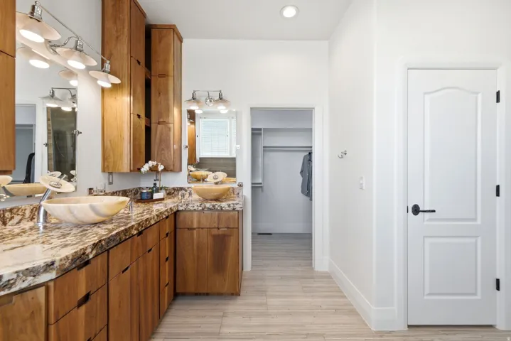 Dual vessel sinks with modern faucets, extensive wood cabinetry, stone-finish countertops, wall-mounted light fixtures, and light-toned flooring