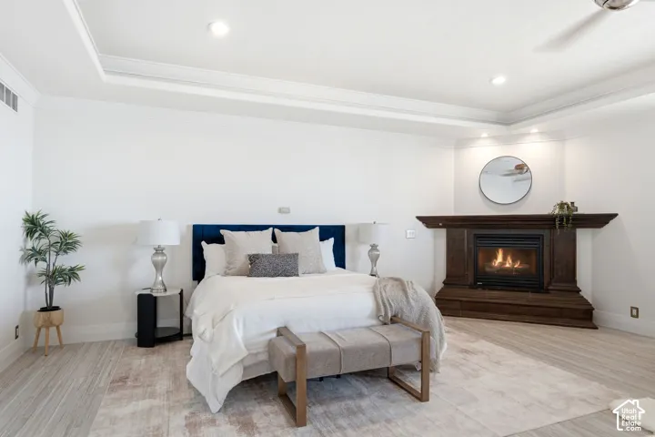 Tray ceiling with recessed lighting, wood-finish flooring, corner fireplace with dark wood surround, and wall-mounted ceiling fan