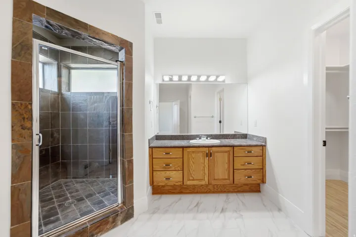 Spacious bathroom featuring a glass-enclosed shower with stone tile, a single vanity with wood cabinetry, a built-in mirror, and white tile flooring