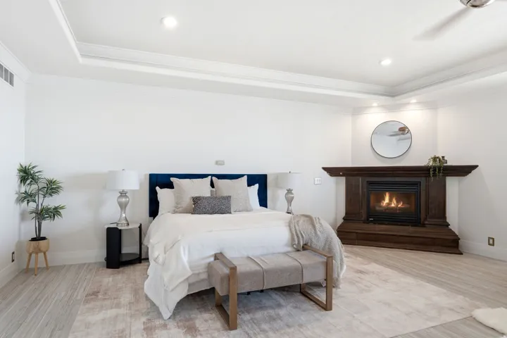 Tray ceiling with recessed lighting, wood-finish flooring, corner fireplace with dark wood surround, and wall-mounted ceiling fan