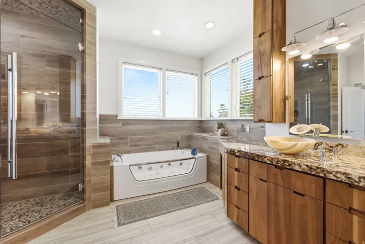 Glass-enclosed shower with tiled walls and pebble floor, soaking tub, wood-finish vanity with stone countertop, and vessel sink
