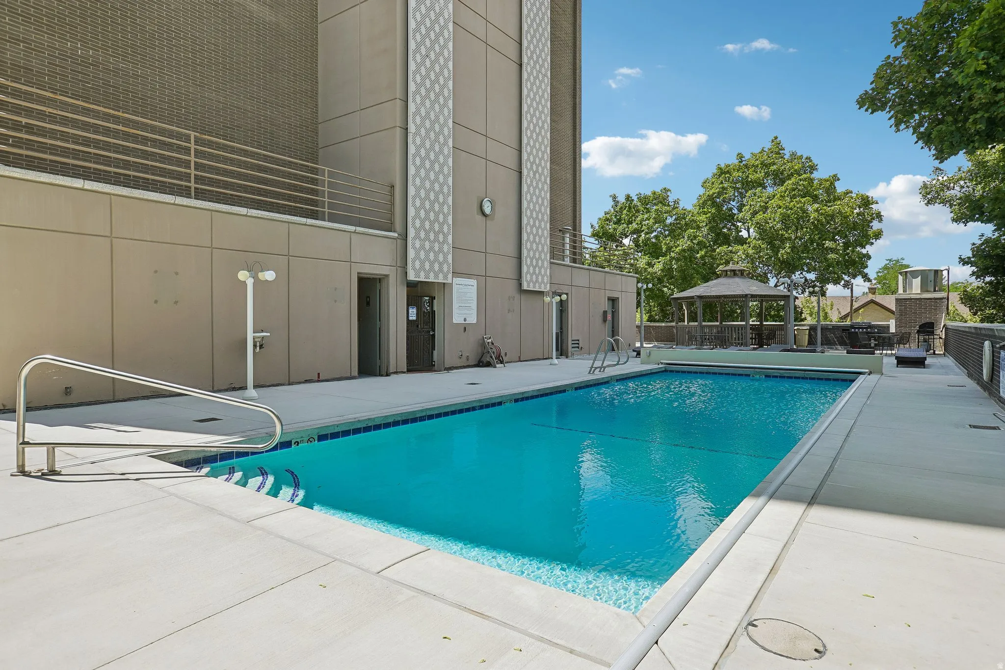 Outdoor swimming pool with integrated steps, surrounded by a concrete deck, featuring a metal gazebo and a low wall with a metal railing