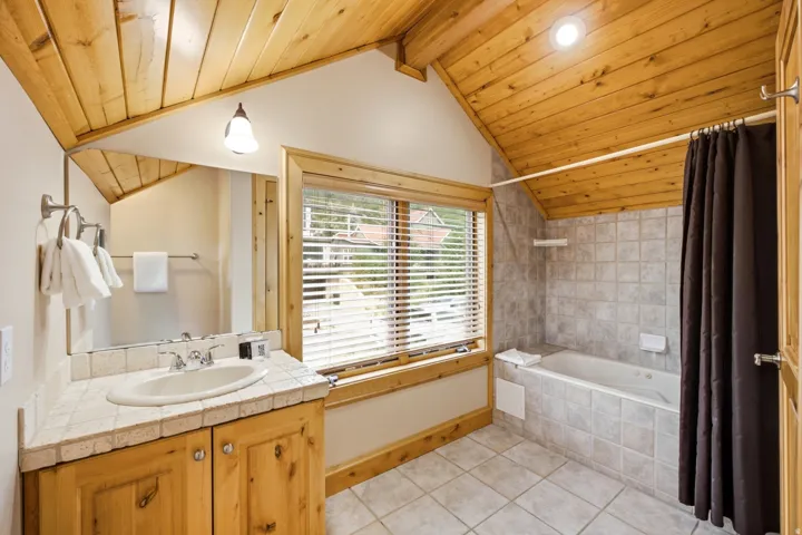 Vaulted wood-paneled ceiling, wood-finish vanity with tiled countertop, built-in soaking tub with tiled surround, and tiled flooring
