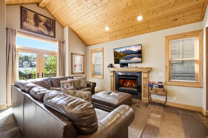 Vaulted wood-paneled ceiling, stone tile flooring, built-in fireplace with a dark stone surround, wood mantel, and French doors leading to an outdoor area