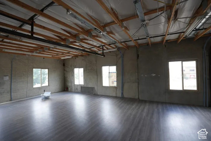 Spacious room featuring wood-finish flooring, exposed ceiling beams, multiple windows, and concrete walls