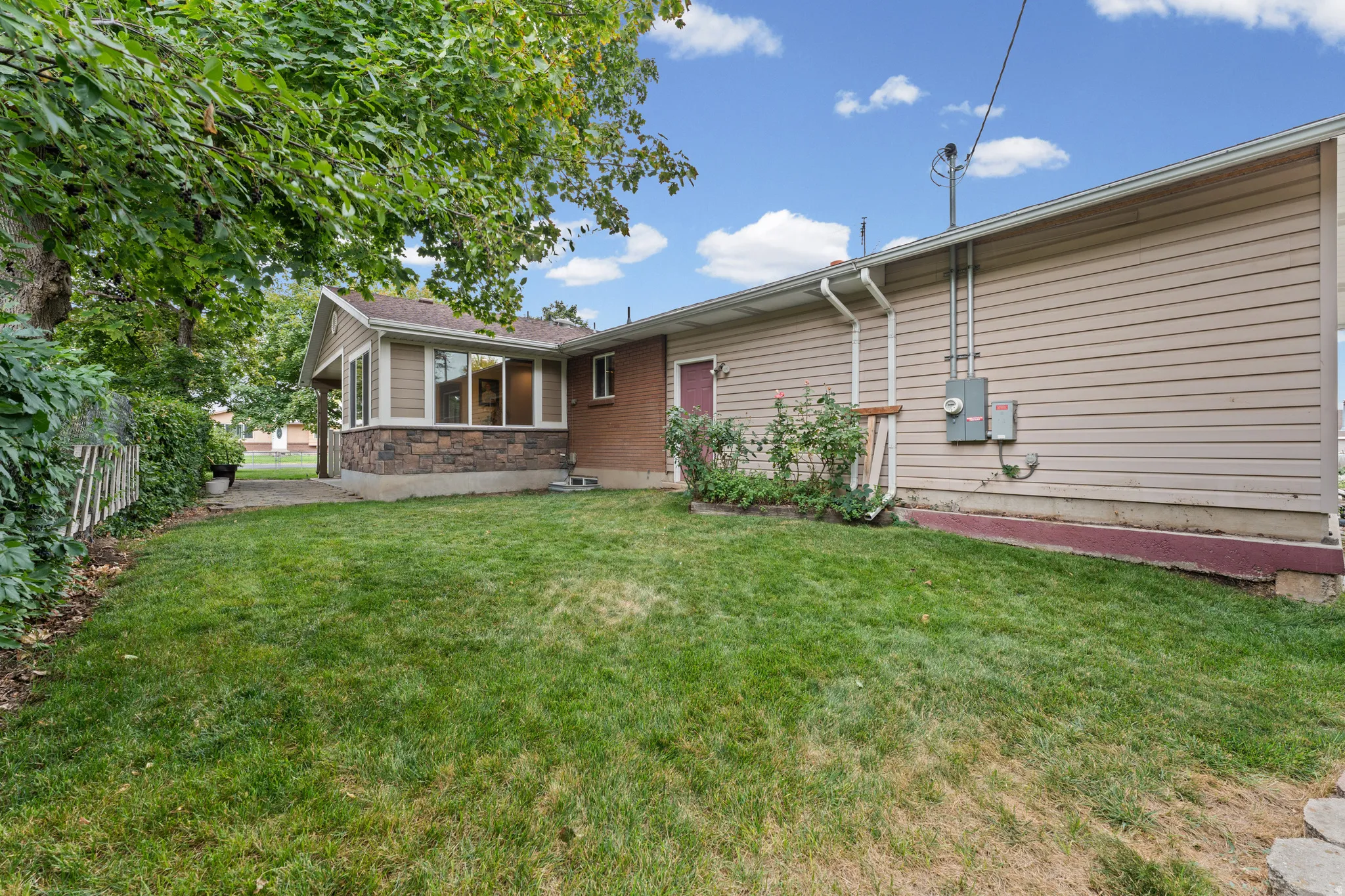 Expansive green lawn with mature perimeter trees, complemented by a stone facade sunroom with multiple windows, and a concrete patio area