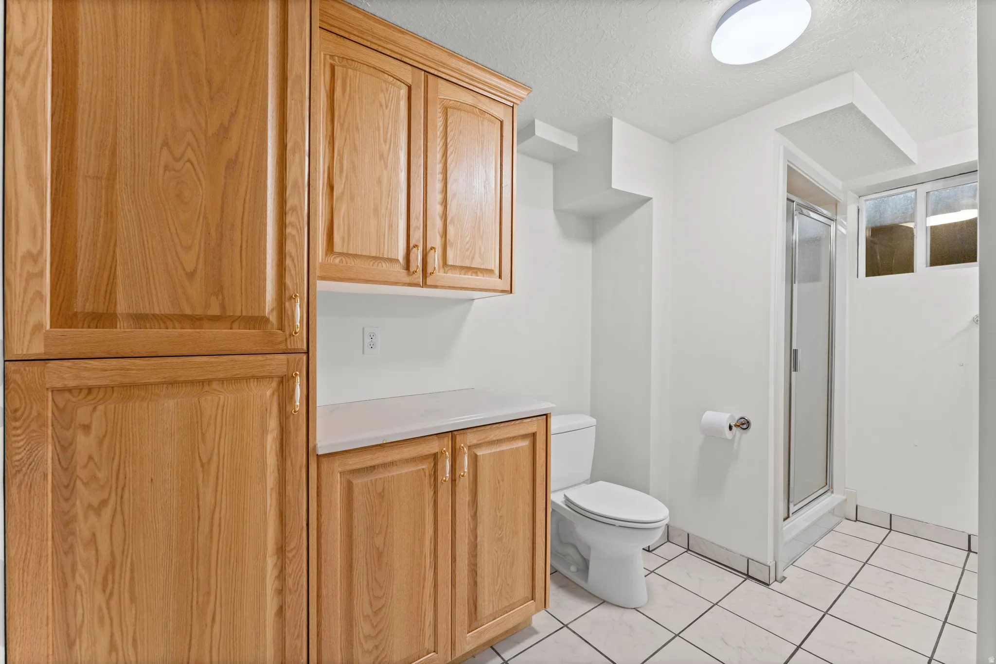 Full-height wood-finish cabinetry with brass hardware, white countertop, white tile flooring, and a glass-enclosed shower