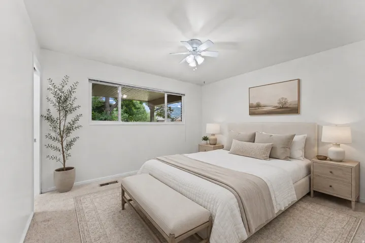 Carpet flooring, white painted walls, window with outdoor scenery, flush mount ceiling fan, and a light-colored headboard