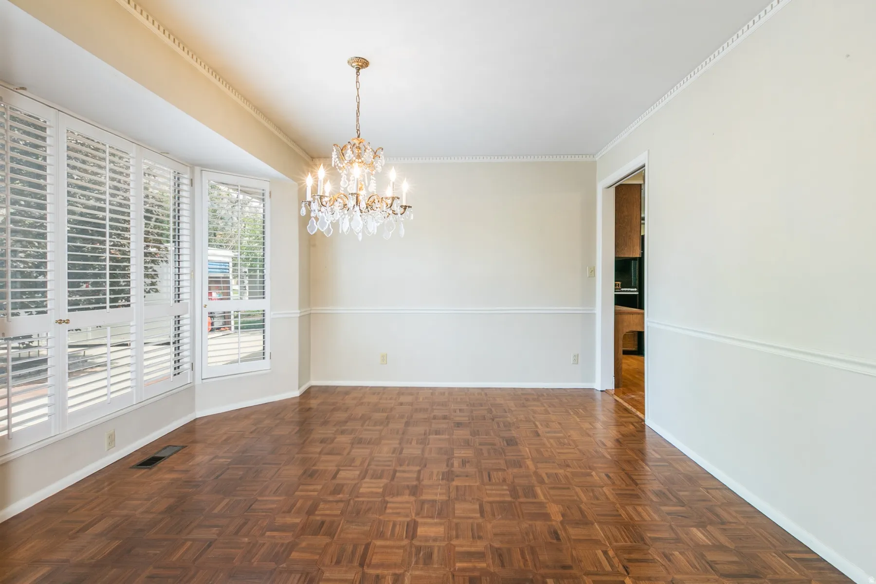 Parquet wood-finish flooring, bay window with plantation shutters, decorative chandelier, crown molding, and chair rail detailing