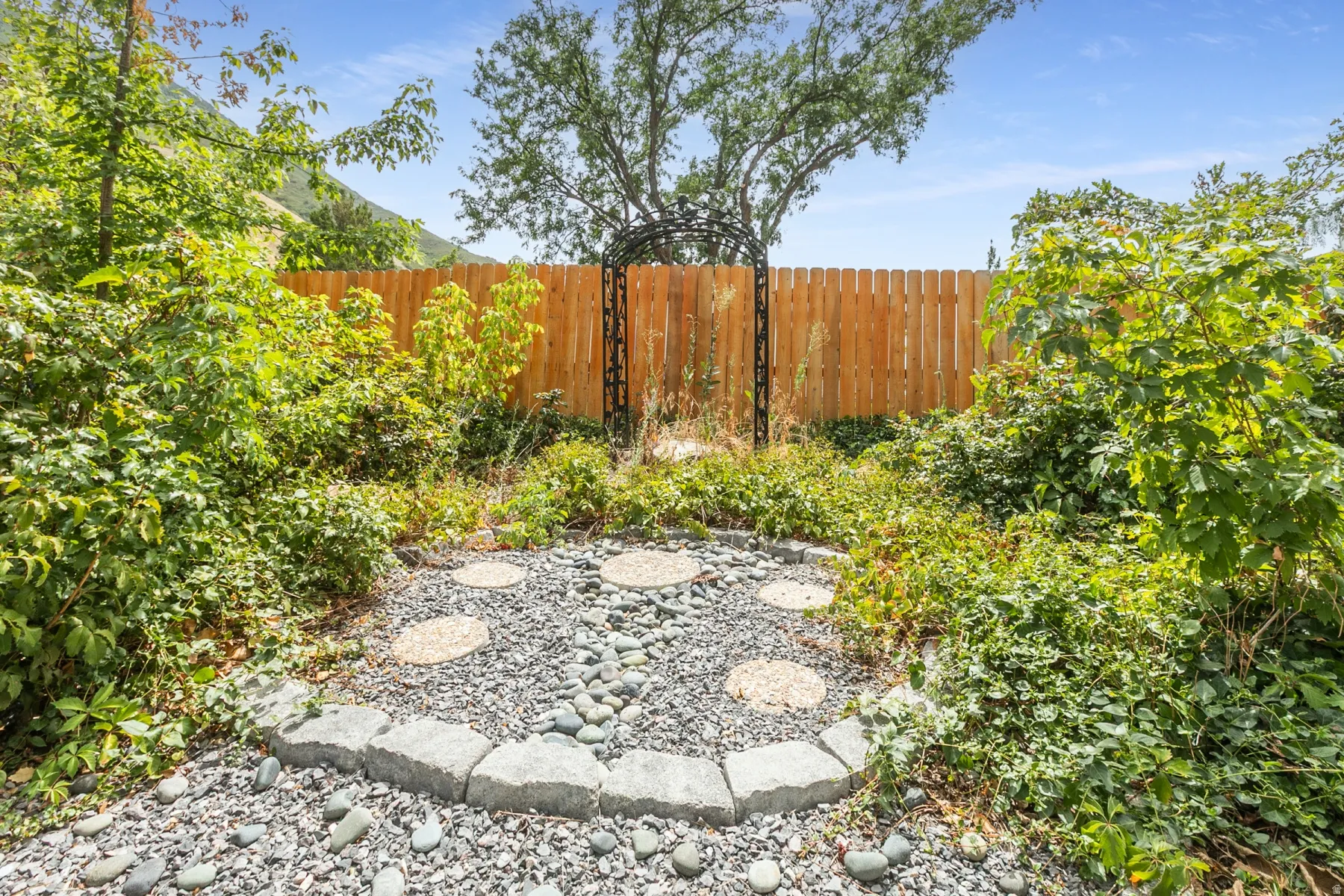 Landscaped garden area featuring a wood fence, decorative metal archway, and a gravel and stone paver path