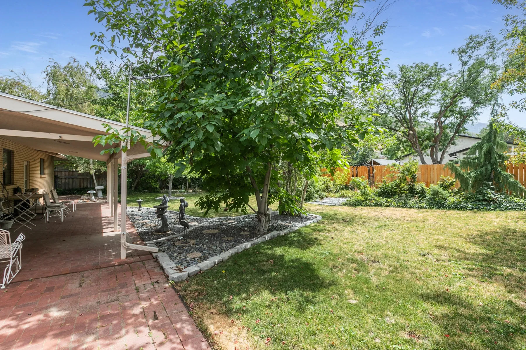 Expansive green lawn with a covered brick patio, mature trees, and a wooden privacy fence
